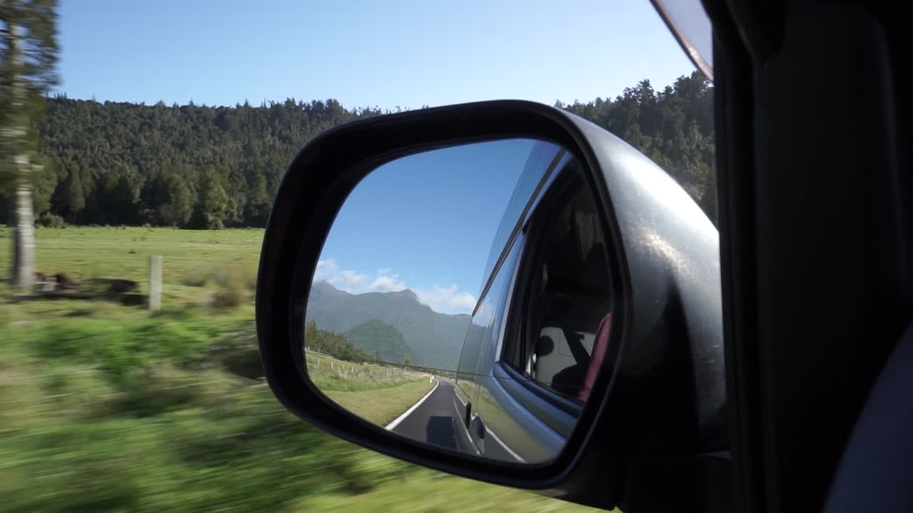 Car driving down the West Coast, New Zealand rear view mirror with mountains, trees and blue sky