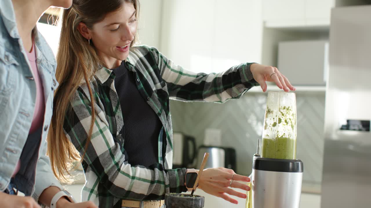 Amiche, donna in plaid che tiene una tazza in mano e la appoggia sul frullatore mentre prepara una bevanda verde per colazione