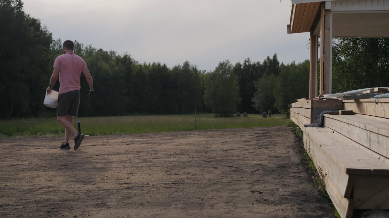 Man walking with a bucket in a rural landscape near a house