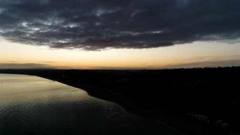 drone overlooking calm shimmering water at sunrise over dark silhouette landscape and clouds