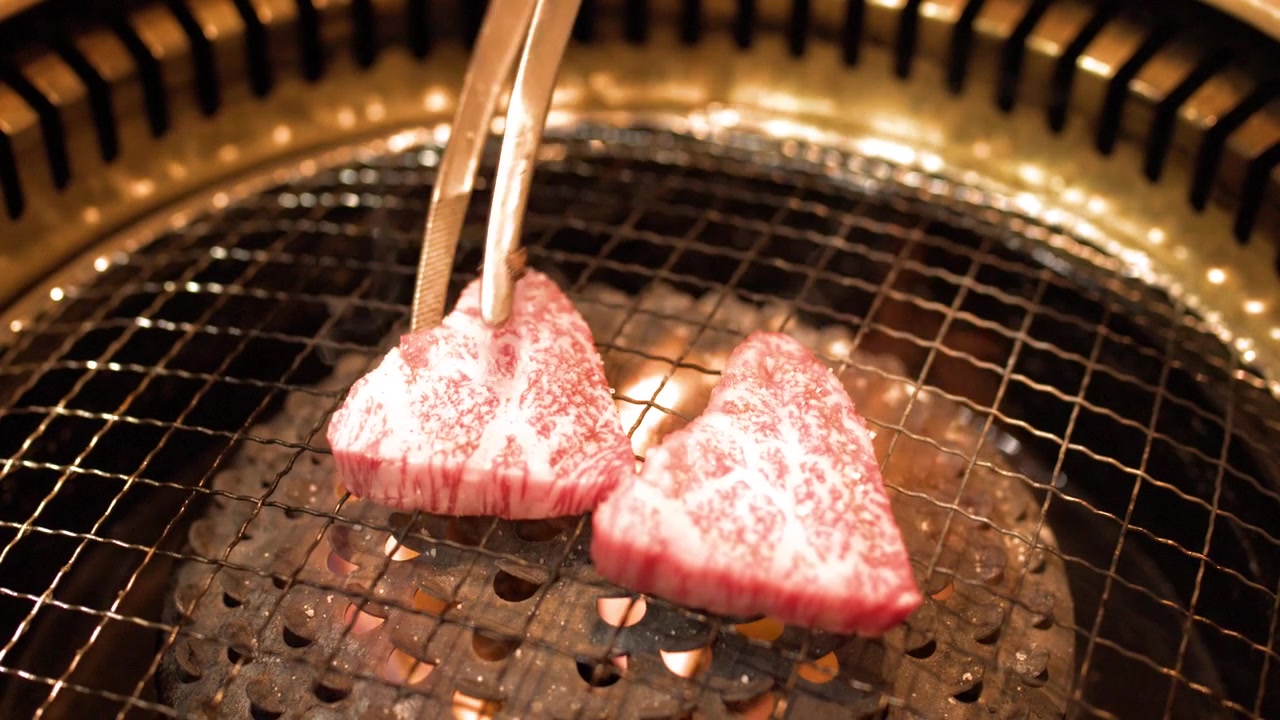 立ち上る煙を伴う熱い炭火グリルに、トングで霜降り牛肉を置くクローズアップ。