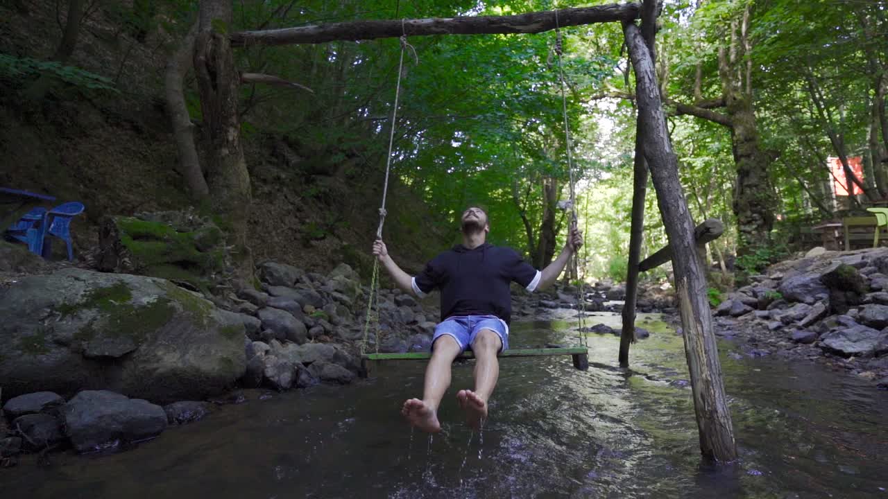 Young man swinging on a swing above the water. Slow Motion.