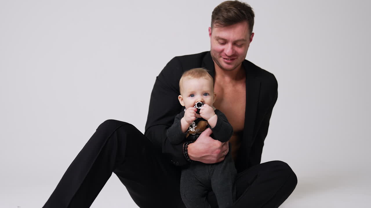 Positive Caucasian man wearing black jacket sits on the floor holding a baby. Proud father with infant son. Studio footage. White backdrop.