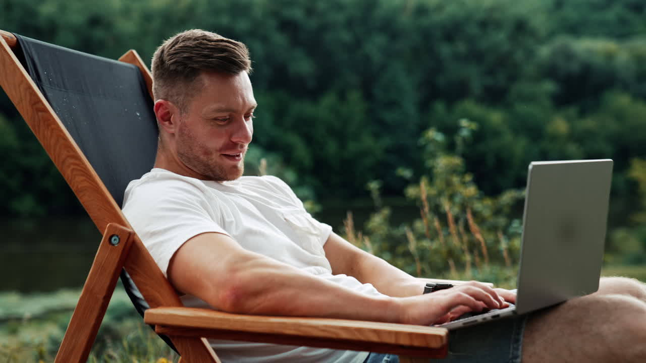 Joyful smiling Caucasian man works on laptop. Freelance businessman working remotely in nature.