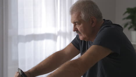 Un-Hombre-De-Mediana-Edad-Está-Haciendo-Ejercicio-Con-Una-Bicicleta-Estática-En-Casa-Para-Mantenerse-En-Forma-Y-En-Buenas-Condiciones-Físicas-Moviéndose-De-Cara-A-Pies