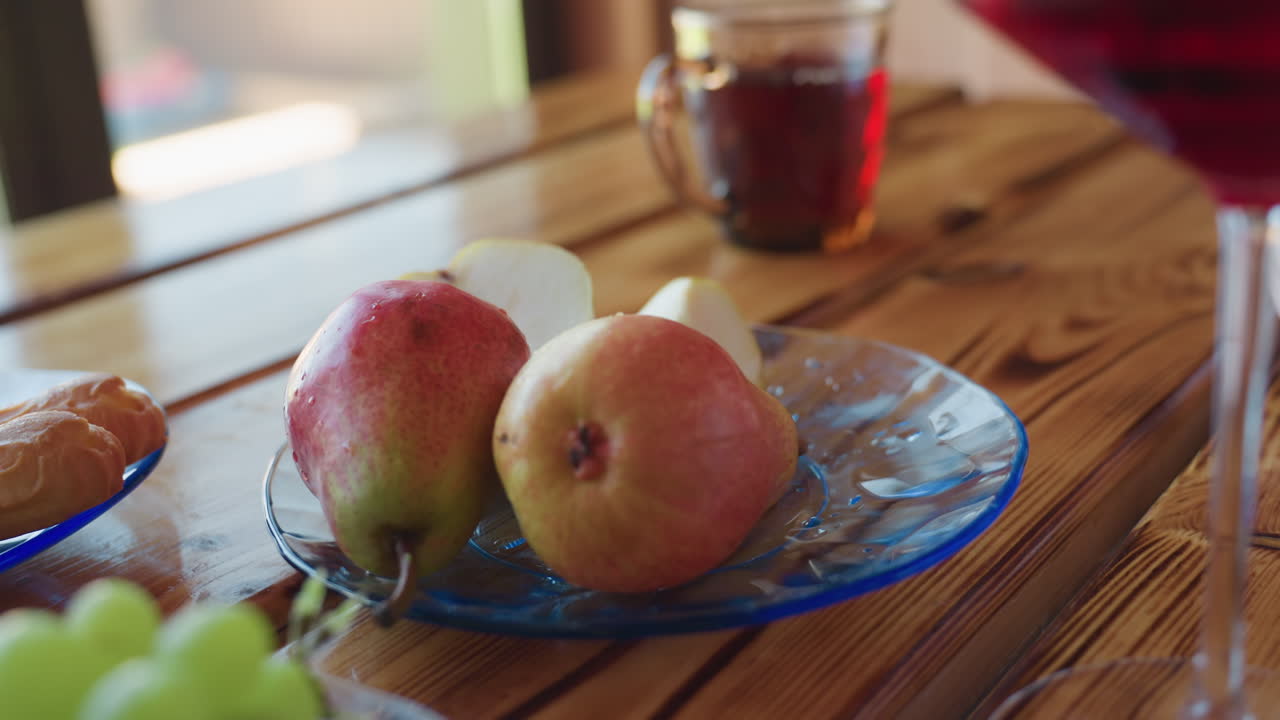 Close up of hand slicing fresh apple with small knife on transparent plate beside ripe pears, wooden table surface and daylight enhance calm indoor food preparation scene with nearby grapes and drink