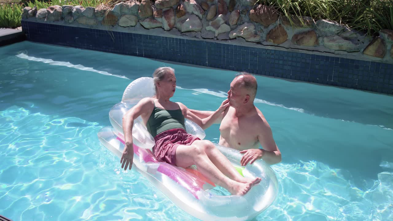 Man is steadying clear float in backyard pool while speaking and touching woman, causing laughter