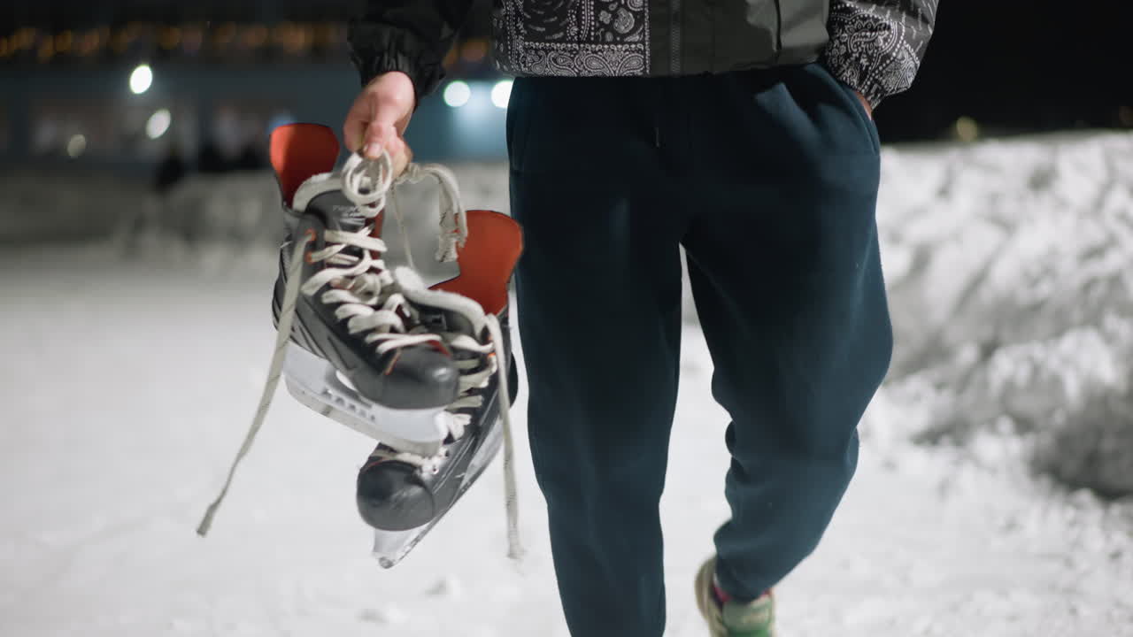 Close up of skater holding skates by laces while walking on snow-covered ground at night, dressed in winter outfit with blurred figures in distant background under soft glowing lights