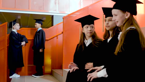 three happy little girls in cap and gown sitting on stairs and saying hello to their classmates at the preschool graduation ceremony