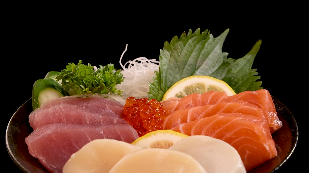 Assorted sashimi and sushi, including salmon, tuna, scallop, and roe, presented on a plate with garnishes, rotating under studio lighting against a black background