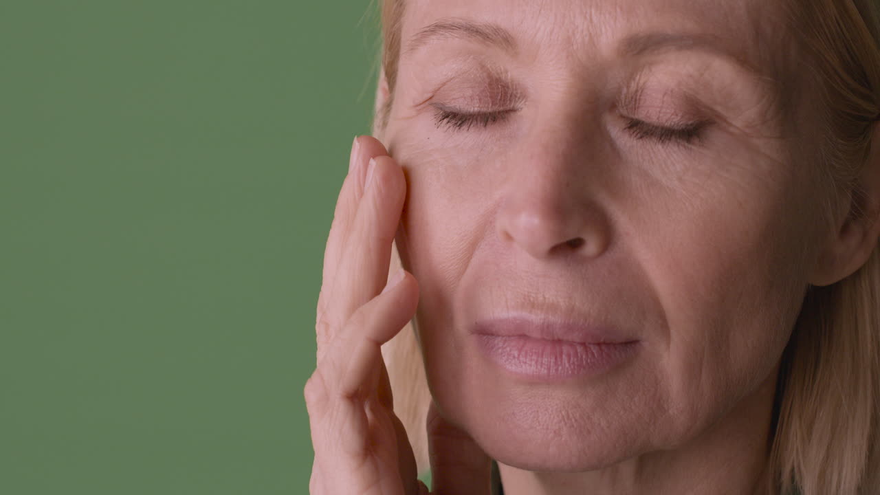 Close Up View Of Blonde Mature Woman With Eyes Closed, Touching Her Face And Looking At Side On Green Background
