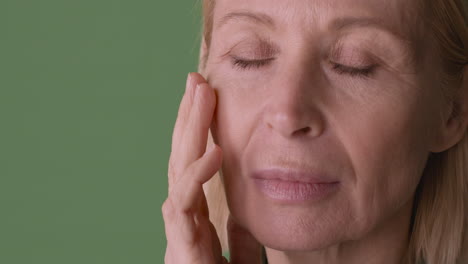 Close-Up-View-Of-Blonde-Mature-Woman-With-Eyes-Closed,-Touching-Her-Face-And-Looking-At-Side-On-Green-Background