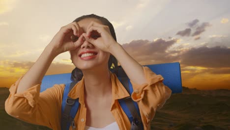 close up of asian female hiker with mountaineering backpack smiling and making binoculars gesture then looking around while standing on the top of mountain during sunset time