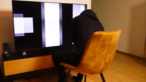 footage of a man in black clothing sitting in front of a big screen television that shows propaganda messages and is brainwashing him