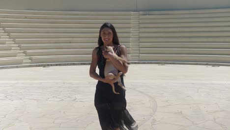 attractive woman in black dress walking in front of amphitheater with kokoni dog on sunny summer day, full frame
