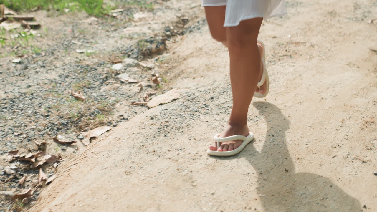 Primo piano di gambe illuminate dal sole che camminano su un sentiero dell'isola, abito bianco che svolazza, infradito che strisciano su ghiaia e sabbia, luce dorata calda che proietta lunghe ombre, passo lento e contemplativo di una viaggiatrice solitaria che esplora una costa tranquilla.