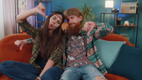 young hippie couple sitting on couch, giving thumbs down