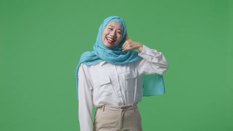 mujer musulmana sonriendo y haciendo llamarme gesto mientras está de pie en la pantalla verde estudio de fondo