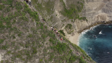 slow pivot around tourists walking along a steep ridge with a pathway to kelingking beach with huge cliff wall in the background, nusa penida, bali, indonesia. aerial shot