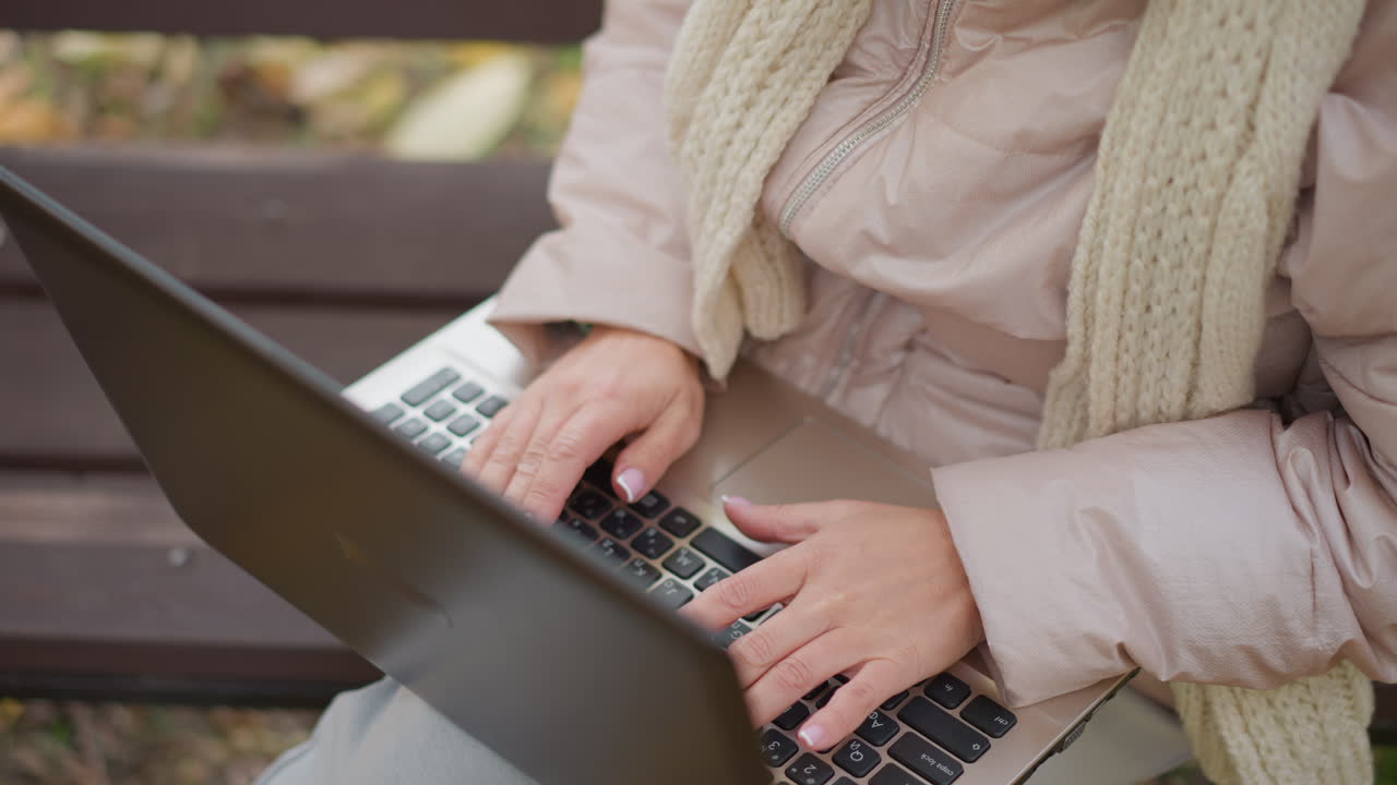 top down view of woman with painted nails typing on laptop while seated on bench in autumn park, finger pointing toward person offscreen, hands moving across keys amid colorful fallen leaves