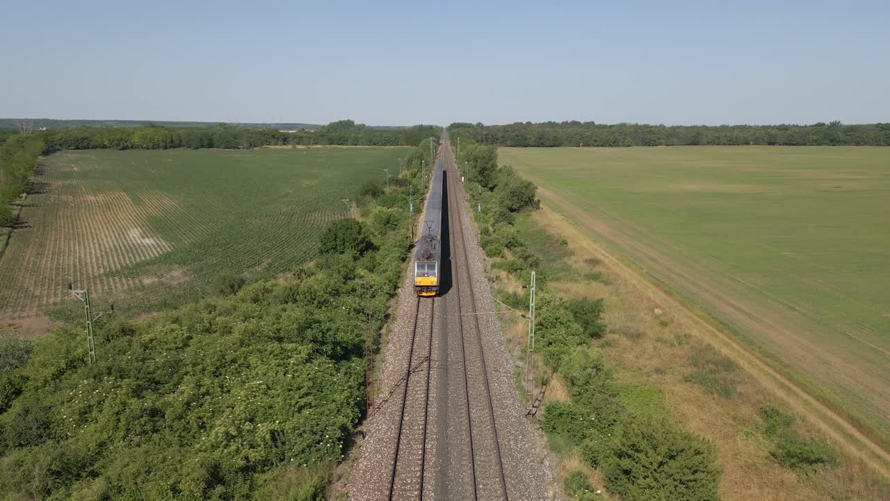 An aerial view of a passenger train moving along railway tracks through wide open countryside. Lush green fields and agricultural land stretch out on both sides under a clear blue sky.