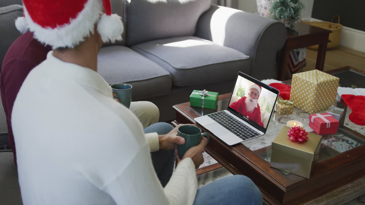 padre y hijo biraciales con sombreros de santa usando una computadora portátil para una videollamada de navidad con santa en la pantalla