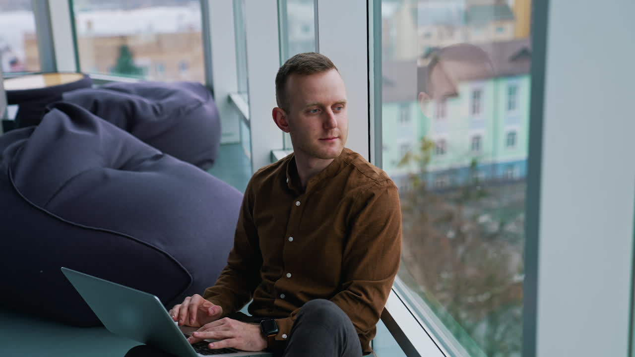 Thoughtful man sit on windowsill in office. Businessman with a laptop on laps looking at cityscape through panoramic window daydreaming about future.