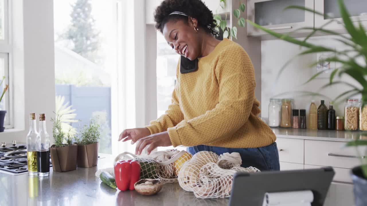 Happy african american woman talking on smartphone unpacking groceries in kitchen, slow motion