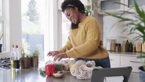 Feliz-Mujer-Afroamericana-Hablando-Por-Teléfono-Inteligente-Desempacando-Comestibles-En-La-Cocina,-Cámara-Lenta