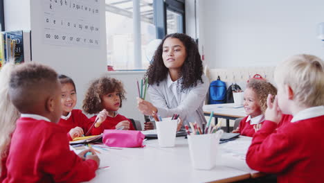 Female-infant-school-teacher-sitting-at-a-table-in-class-giving-pencils-to-children