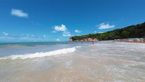 fotografía de mano de pequeñas olas que chocan contra el famoso destino turístico tropical de la playa de madeiro en pipa, brasil, con turistas nadando y descansando bajo paraguas rodeados de enormes acantilados