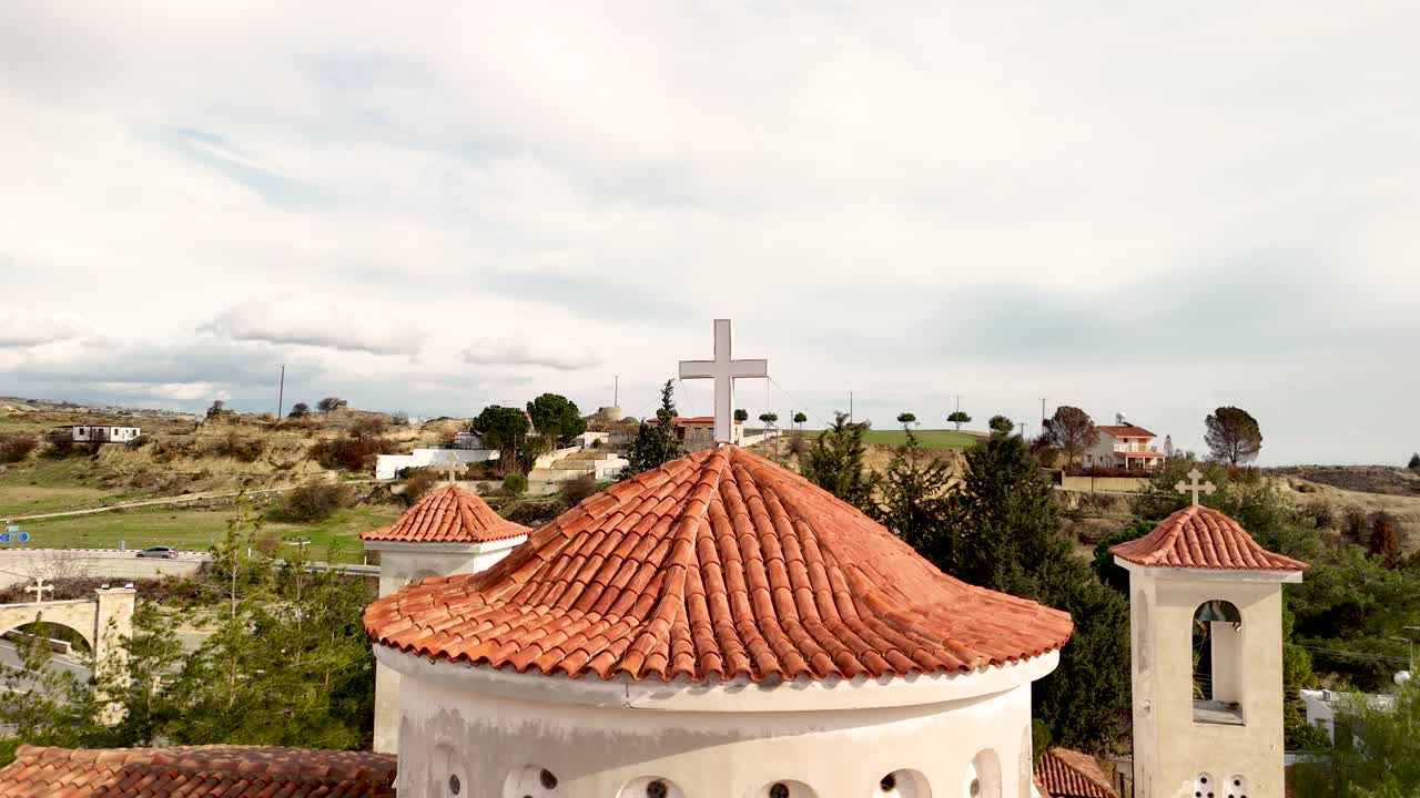 Una vista di una chiesa con una grande croce in cima. La chiesa si trova in un villaggio con molte case. Le colline sono vicine e le nuvole coprono parti del cielo