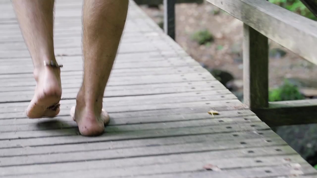 Kamera w zbliżeniu śledzi stopy mężczyzny idącego po drewnianym moście i wchodzącego głębiej na leśną ścieżkę.