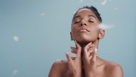 retrato de belleza en cámara lenta mujer afroamericana atractiva plumas cayendo sobre la piel lisa tocando hombros desnudos disfrutando del cuidado natural de la piel acariciando suavemente el cuerpo en fondo azul
