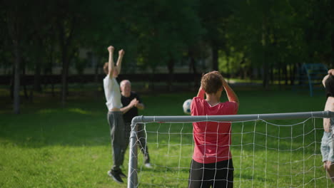 Family engages in a lively outdoor soccer game. The father runs with his face covered, the young son jumps in joy, and the son in red holds his head as a ball flies through the air