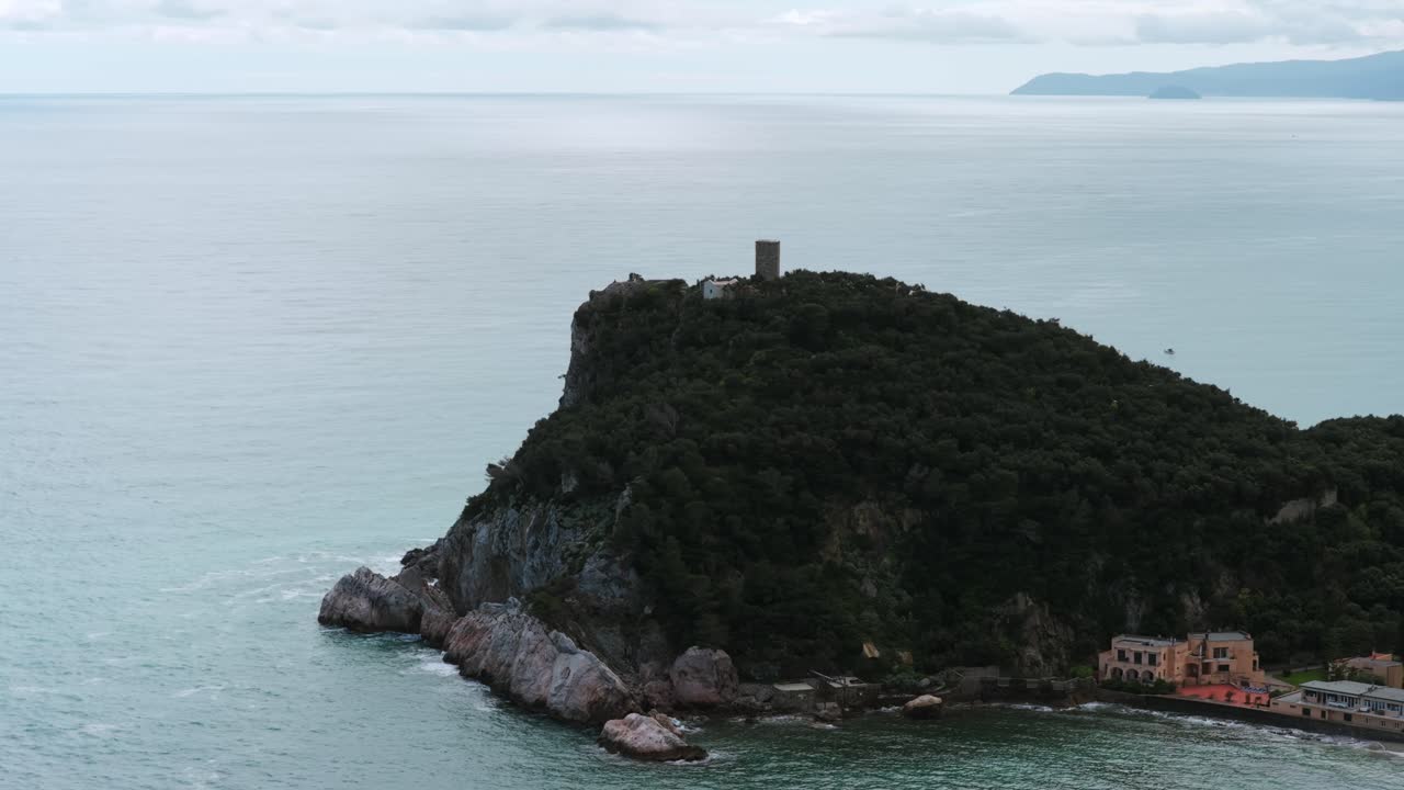 Drone footage showcasing a rugged, vegetated headland extending into the clear blue Mediterranean Sea near the coastal town of Noli, Liguria, Italy