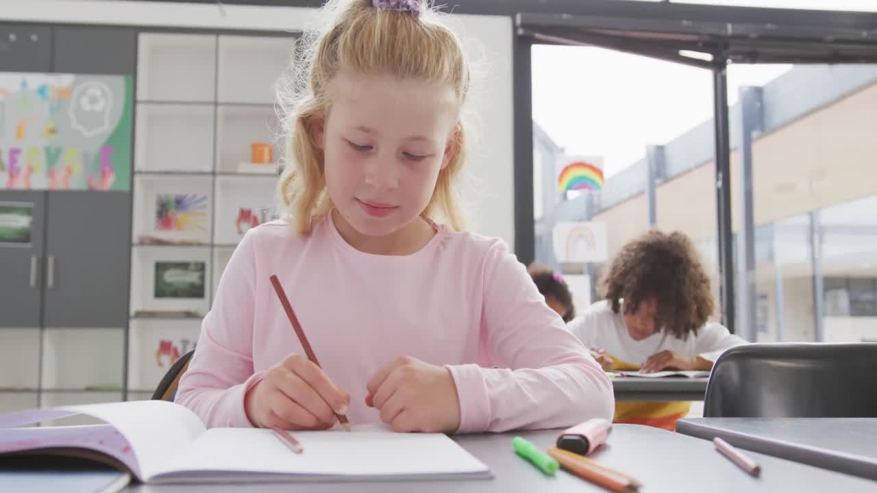 Video van een blij kaukasisch schoolmeisje dat aan een bureau schrijft in een diverse schoolklas.