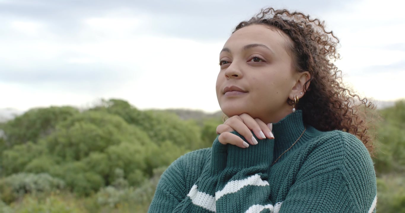 Donna afroamericana adulta con maglione verde, con le mani giunte, che sorride sulle colline.