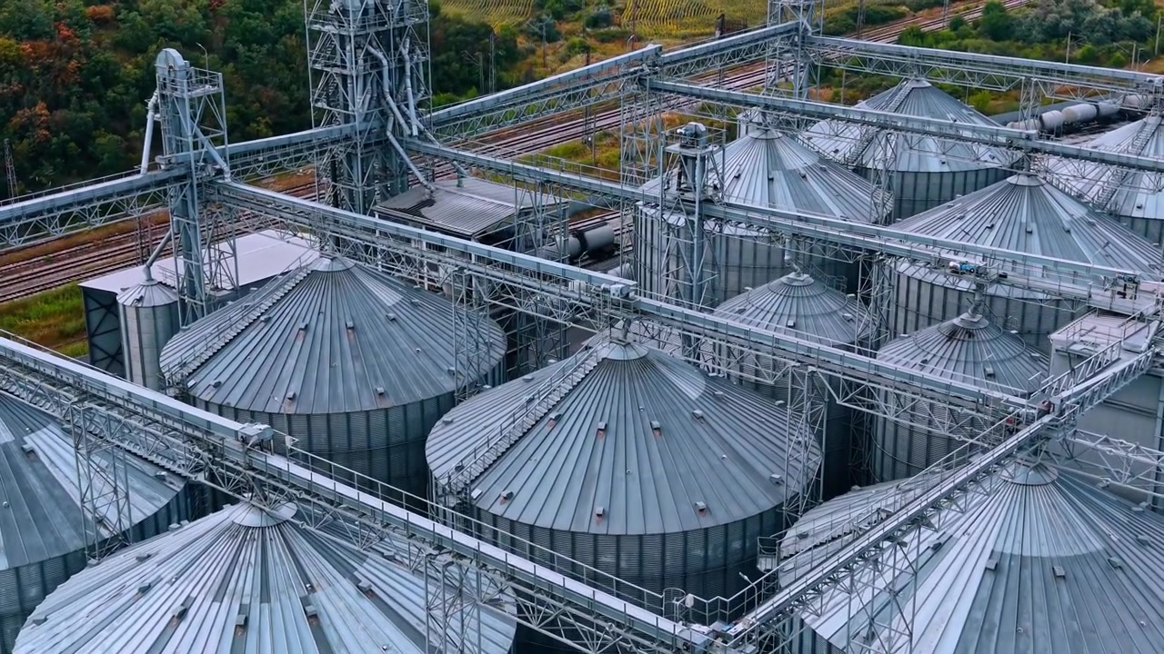 Vista aérea de telhados e treliças de silos de aço. Estrutura metálica se estende sobre silos de armazenamento circulares perto de linhas férreas à luz do dia.