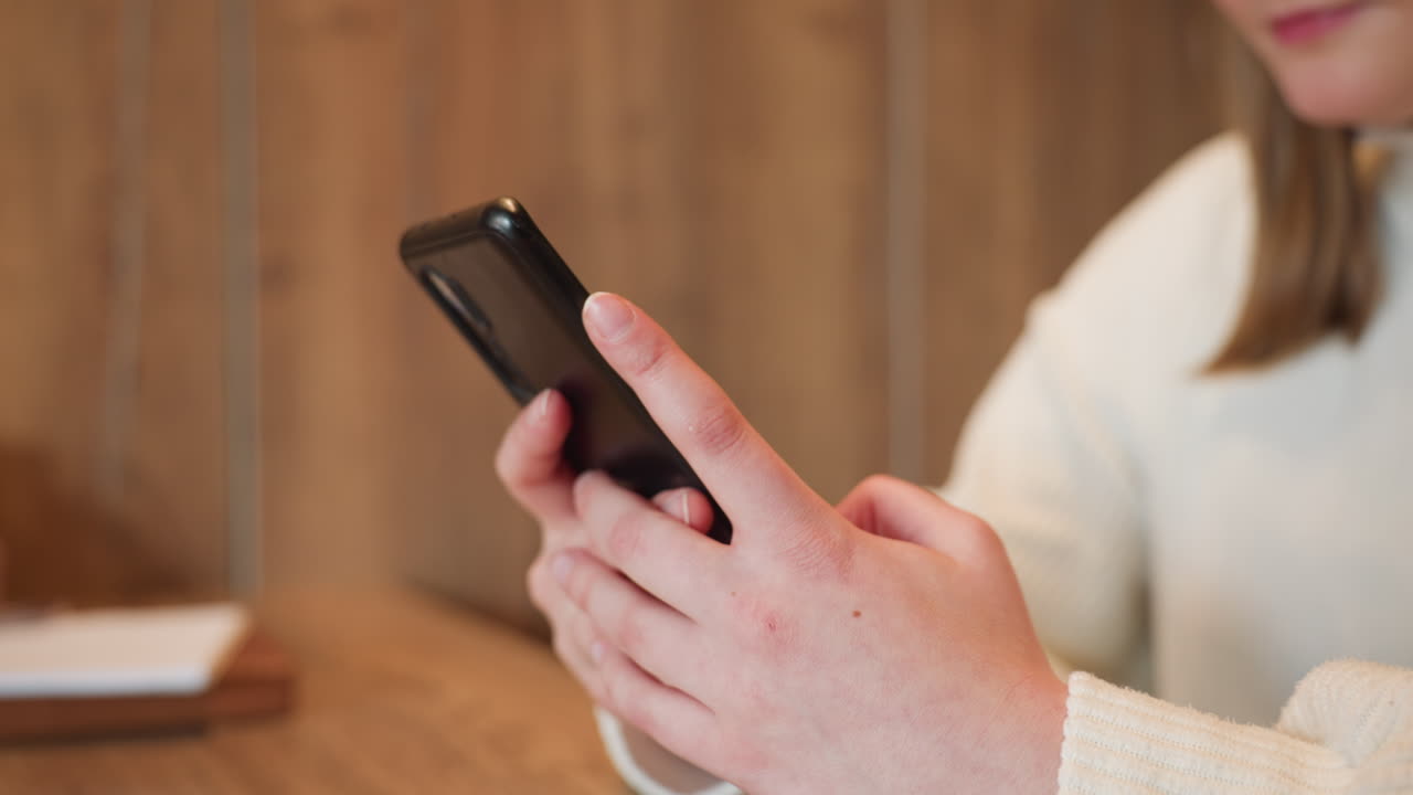 Close up hand view of lady operating phone while seated in cozy cafe with warm wooden background, fingers interacting with screen in calm relaxed moment of digital communication and connection