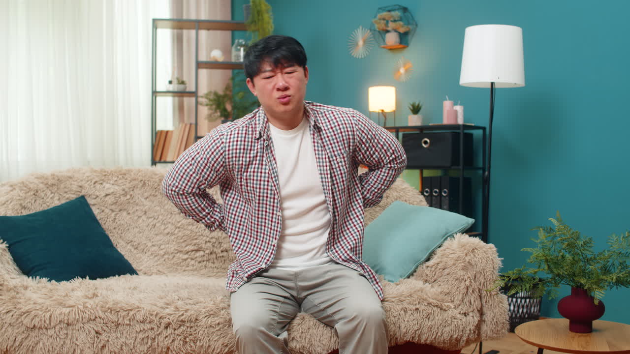 Asian man grimacing from strong back pain carefully sitting down painful strain discomfort tension