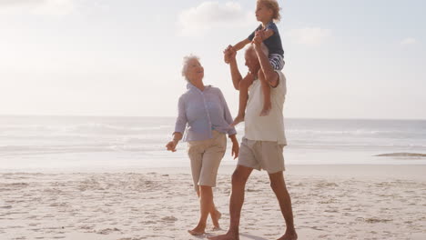 Grandparents-Carrying-Grandson-On-Shoulders-As-They-Walk-On-Beach
