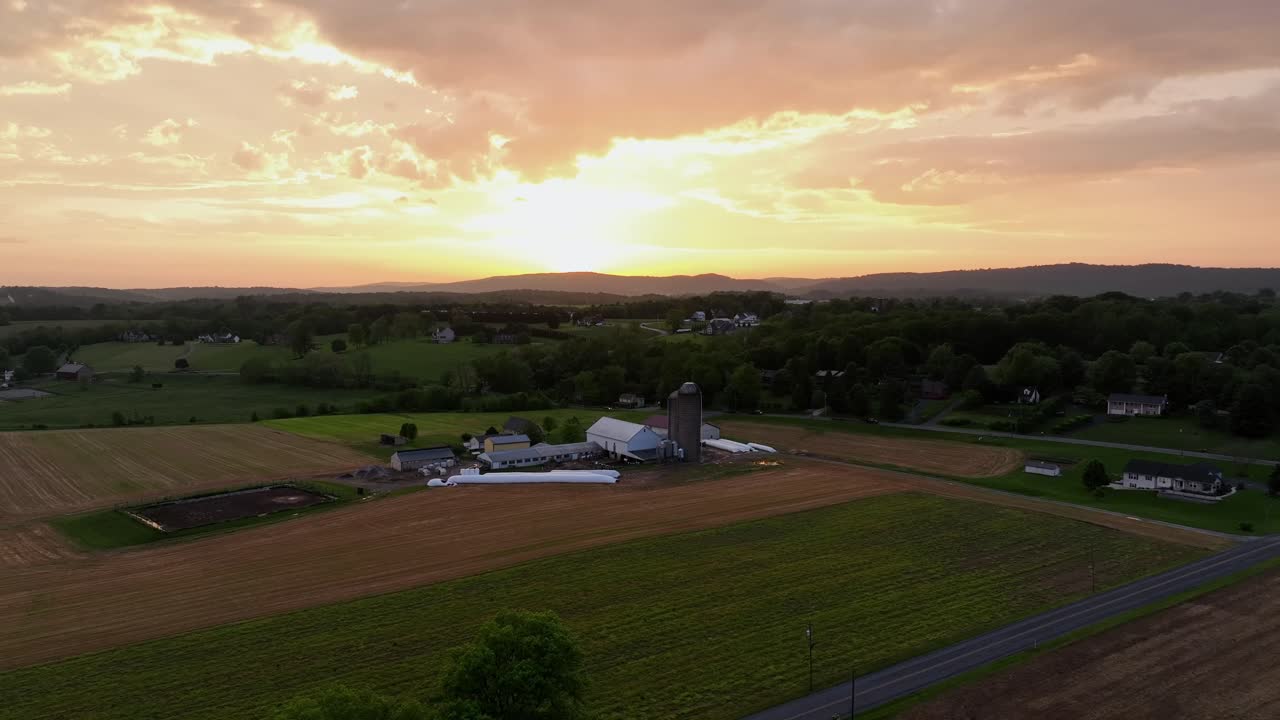 Fattoria americana con silo di stoccaggio e campi colorati con tramonto dorato dietro le nuvole. Ampia ripresa aerea in risalita all'indietro. Paesaggio rurale calmo e pacifico negli Stati Uniti d'America.