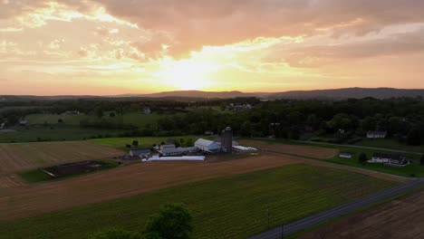 american farm with silo storage and colored fields with golden sunset behind clouds. aerial backwards rising wide shot. rural calm and peaceful landscape in united states of america.