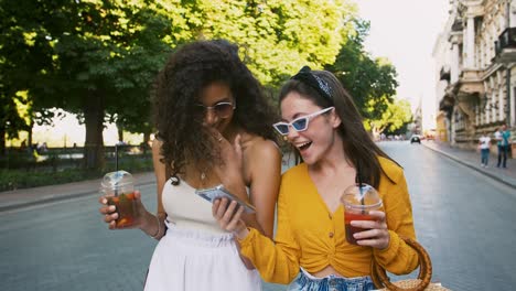 two young ladies are holding cold drinks in plastic cups. laughing, looking surprised while watching video on cell phone. walking along city avenue