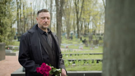 hombre con impermeable negro y traje sosteniendo rosas rojas frente a una lápida en un cementerio 1