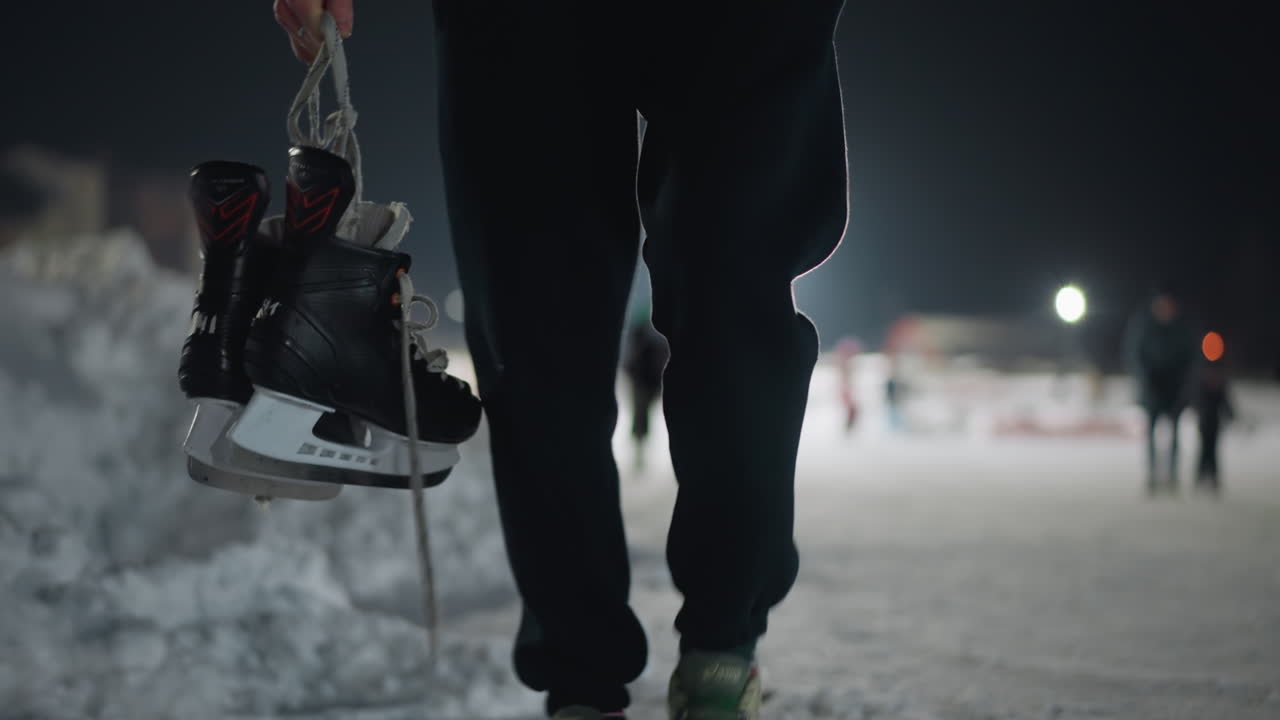 low angle close up of skater walking on snow-covered path holding black ice skate by lace, with cold night backdrop and soft lights creating serene winter atmosphere