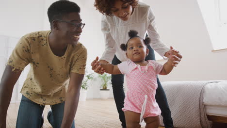 Una-Niña-Aprende-A-Caminar-En-Casa-Mientras-Los-Padres-Se-Sientan-En-El-Suelo-Y-La-Alientan