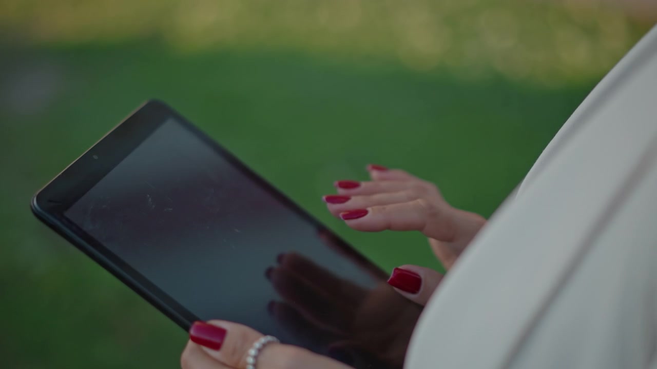 Close-up of hands holding tablet outdoors while fingers tap and swipe blank touchscreen, showing red manicured nails, decorative ring, and soft natural light over blurred green background