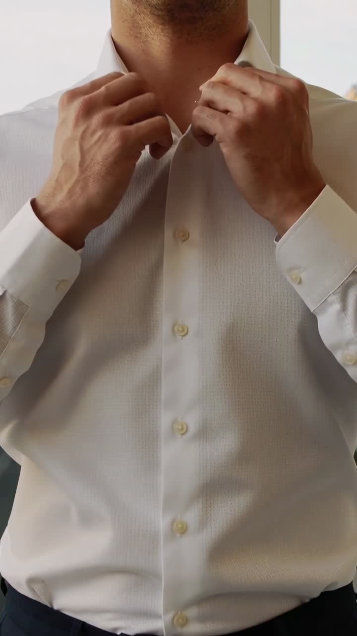 Video verticale: Preparandosi per una riunione, un uomo sistema il colletto della camicia bianca vicino alla finestra, lisciando il bottone.